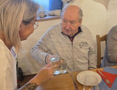 An einem gedeckten Holztisch stoßen eine Dame aus der Frauenbewegung und ein Bewohner mit Gläsern an, während vor ihnen Teller, Besteck, Weingläser und eine dekorative Kerze auf einer Baumscheibe stehen.