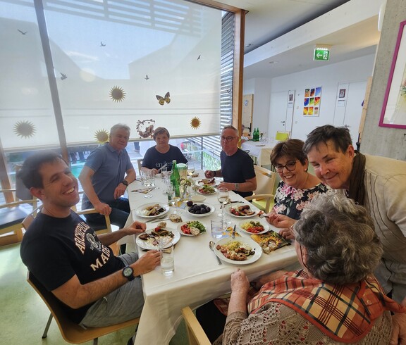 Ein großer Tisch voller Bewohnerinnen und Bewohner, Angehöriger und Mitarbeitender bildet beim Sommerfest im Pflegewohnhaus den Mittelpunkt eines gemeinsamen Essens in freundlicher und festlicher Umgebung.