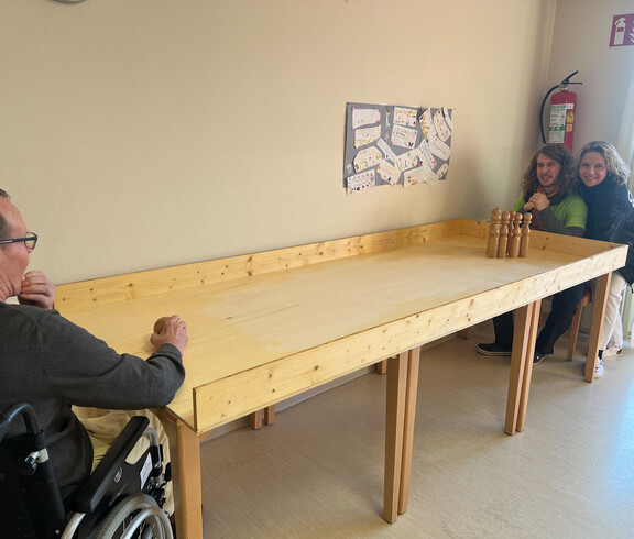 Bewohner stößt eine Kugel über die Holzbahn, zwei Pflegekräfte stehen am anderen Ende und beobachten das Geschehen.