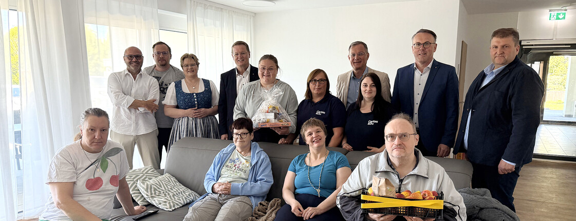Mehrere Personen sitzen in einem Raum auf einem Sofa, dahinter stehen noch weitere Personen.