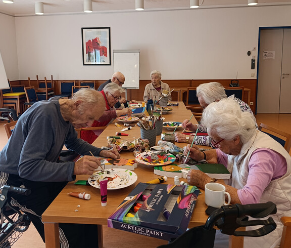 Mehrere Personen sitzen am Tisch und gestalten Krippenfiguren mit Farben