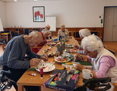 Mehrere Personen sitzen am Tisch und gestalten Krippenfiguren mit Farben