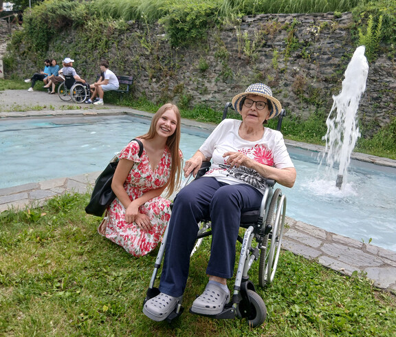 Bewohnerin im Rollstuhl und Schülerin verbringen Zeit miteinander an einem Brunnen im Park.