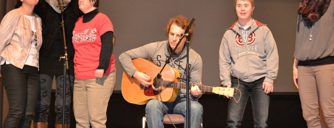 Ein Musiker mittleren Alters spielt auf der Bühne eine akustische Gitarre. Ihn umgeben Menschen mit Behinderung.