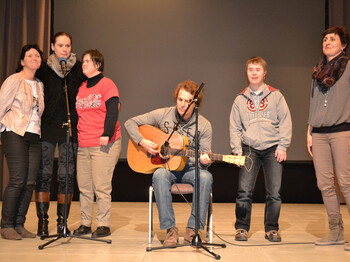 Ein Musiker mittleren Alters spielt auf der Bühne eine akustische Gitarre. Ihn umgeben Menschen mit Behinderung.
