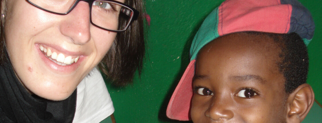 Erwachsene, junge Frau mit Brille, weißem t-Shirt und einem schwarzen Schal trifft sich mit einem kleinen Jungen, der eine Schildkappe falsch auf seinem Kopf hat. Beide strahlen vor Freude. 