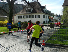 Ein Lauf um Wohnhäuser im Freien. Die Menschen tragen Sportkleidung und laufen auf einem geteerten Boden. 