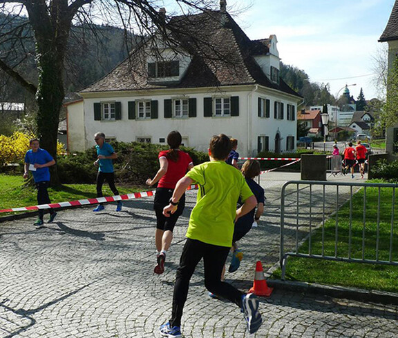 Ein Lauf um Wohnhäuser im Freien. Die Menschen tragen Sportkleidung und laufen auf einem geteerten Boden. 