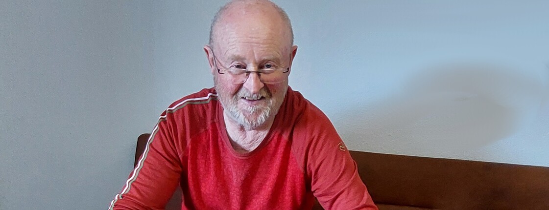 Älterer Herr im roten langärmligen T-Shirt sitzt beim Tisch mit einem Zettel, Taschenrechner, Lineal und Bleistift