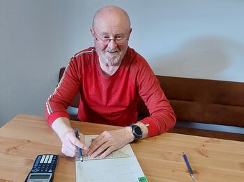 Älterer Herr im roten langärmligen T-Shirt sitzt beim Tisch mit einem Zettel, Taschenrechner, Lineal und Bleistift