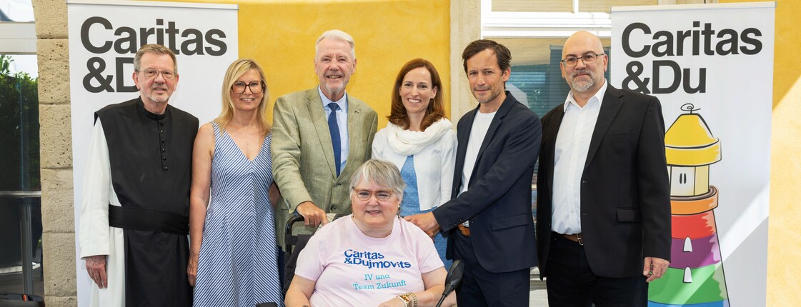 Gruppenfoto vor zwei „Caritas & Du“-Bannern: Mehrere Personen stehen zusammen, in der Mitte sitzt eine Person im Rollstuhl 