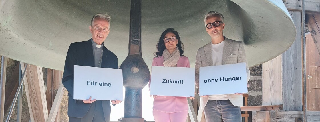2 Männer und eine Frau stehen vor einer großen Kirchenglocke