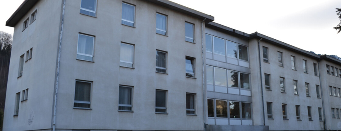 An einer Straße steht ein dreistöckiges größeres Gebäude, das weder alt noch neu in seiner Erscheinung ist. Die Aussenfassade ist weiß. Vor dem Block stehen etliche Autos in einer Reihe auf Parkplätzen. 