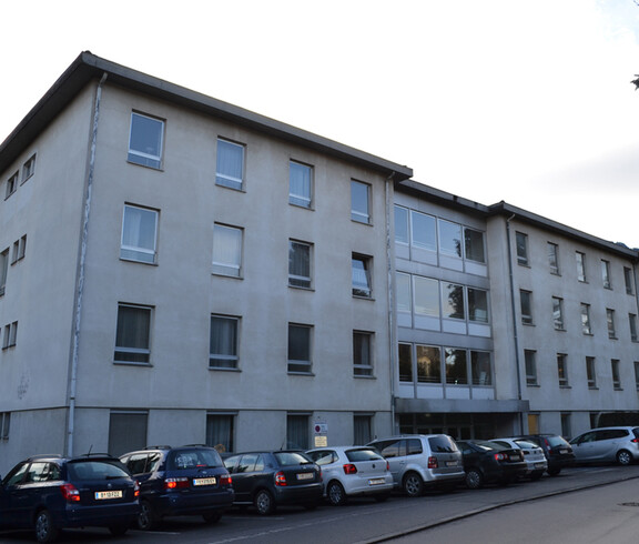 An einer Straße steht ein dreistöckiges größeres Gebäude, das weder alt noch neu in seiner Erscheinung ist. Die Aussenfassade ist weiß. Vor dem Block stehen etliche Autos in einer Reihe auf Parkplätzen. 