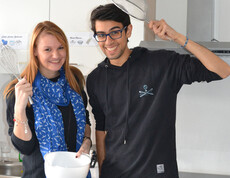 Zwei junge Erwachsene, eine Frau und ein Mann, stehen in einer Küche. Sie haben dabei Kochutensilien wie ein Sieb und eine Schüssel in der Hand. Beide sind dunkel gekleidet, die Frau trägt zusätzlich einen blauen Schal und der Mann eine Brille. 