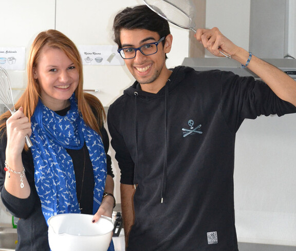 Zwei junge Erwachsene, eine Frau und ein Mann, stehen in einer Küche. Sie haben dabei Kochutensilien wie ein Sieb und eine Schüssel in der Hand. Beide sind dunkel gekleidet, die Frau trägt zusätzlich einen blauen Schal und der Mann eine Brille. 