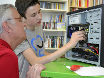 Generationenaustausch: Ein junger Bursche mit grauem t-Shirt und ein älterer Mann mit rotem Polo reparieren einen Computer. Die jeweiligen Hardwareteile sind dabei gut ersichtlich. Der PC steht auf einem grünen Tisch. 