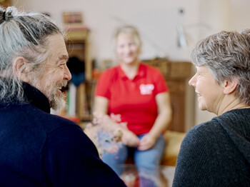 Ein Mann und eine Frau lächeln sich an, eine Caritas Pflegekraft im Hintergrund