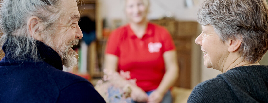 Ein Mann und eine Frau lächeln sich an, eine Caritas Pflegekraft im Hintergrund