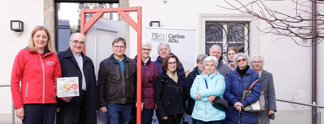 Caritas Steiermark Direktorin Nora Tödtling-Musenbichler steht mit Diözesanbischof Wilhelm Krautwaschl und einer Gruppe von Haussamler:innen im Innenhof der Stadtpfarrkirche Graz um einen geöffneten Türrahmen zusammen.