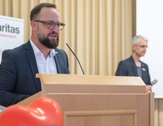 Mann mit dunklen kurzen Haaren, Bart und Brille steht am Rednerpult und spricht ins Mikrofon.