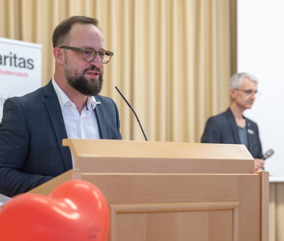 Mann mit dunklen kurzen Haaren, Bart und Brille steht am Rednerpult und spricht ins Mikrofon.