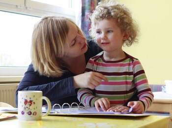 Ein kleines Kind steht an einem Tisch. Vor ihm liegt eine aufgeschlagene Ringmappe. Neben ihm sitzt eine Frau, die sich von der Seite zu ihm rüberbeugt.