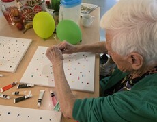 Eine Bewohnerin des Pflegewohnhauses gestaltet konzentriert mit bunten Farbtupfern eine Leinwand und nimmt aktiv am kreativen Malangebot teil.