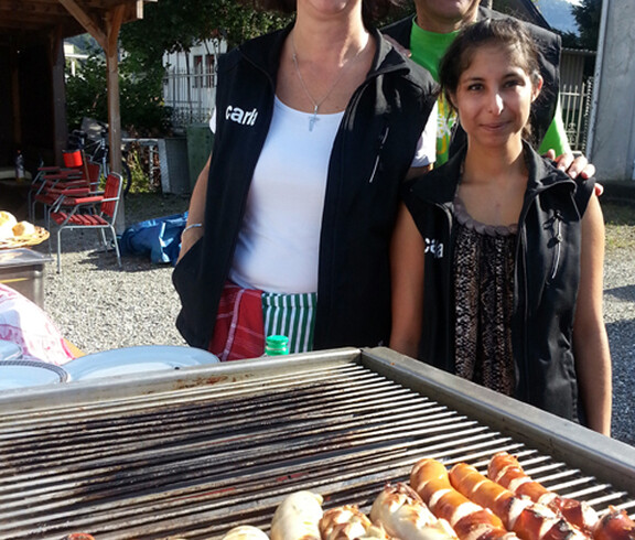 An einem Grill, auf dem sich saftige Würste befinden, stehen drei carla Mitarbeiter.