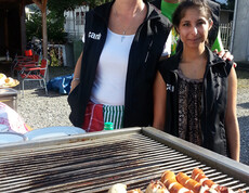 An einem Grill, auf dem sich saftige Würste befinden, stehen drei carla Mitarbeiter.