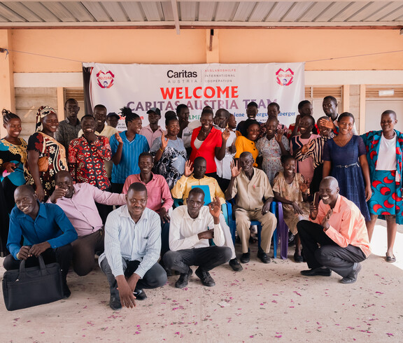 Viele Erwachsene aus dem Südsudan stehen vor der Schule in Gumbo vor einem Plakat, auf dem steht "Welcome Caritas Austria Team".