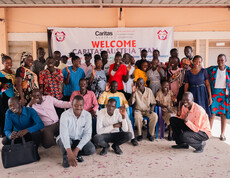 Viele Erwachsene aus dem Südsudan stehen vor der Schule in Gumbo vor einem Plakat, auf dem steht "Welcome Caritas Austria Team".
