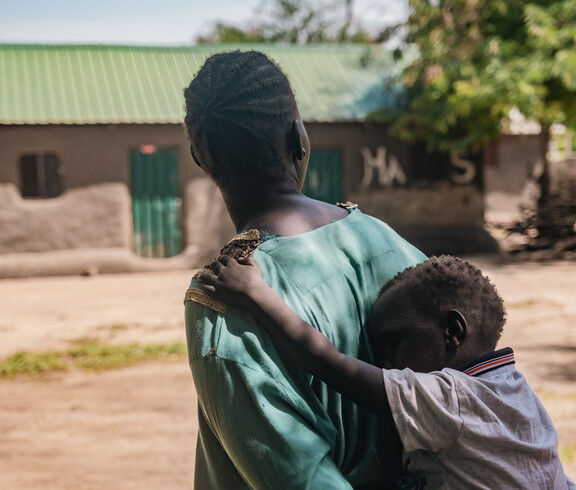 Eine Schwarze Frau trägt ein kleines Kind auf ihrem Rücken. Im Hintergrund ist ein Lehmhaus zu sehen.