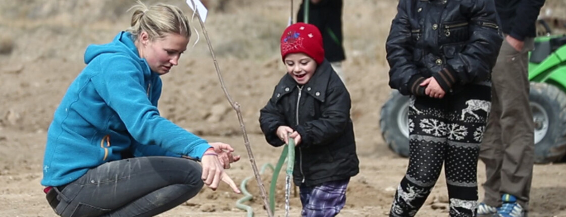 Zwei armenische Kinder planzen mit Hilfe einer Freiwilligen einen Baum in der Landschaft. Alle sind dabei in warmen Winterklamotten gehüllt und dunkel bekleidet. Die junge Frewillige trägt im Kontrast zu den anderen drei sich auf dem Foto befindenen Personen einen hellblauen Sweater. Die Kinder sind angesichts der Situation und der Aufmerksamkeit, die sie erfahren, erfreut. 