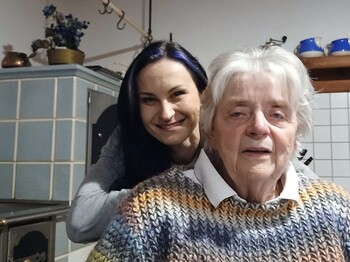 Im Vordergrund ist eine ältere sitzende Dame zu sehen. Dahinter beugt sich eine junge Frau mit langen dunklen Haaren runter und lächelt in die Kamera. 
