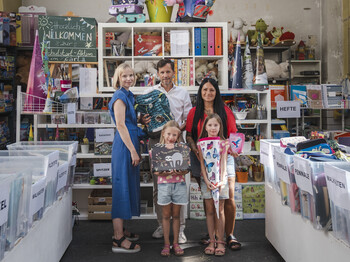 Mehrere Personen stehen in einem Raum mit Regalen und Kisten voller Schulmaterialien. Die Kinder halten Schultüten und Schulzubehör, während rundherum sortierte Hefte, Mappen, Federpennale und weitere Schulsachen für den Schulstart vorbereitet sind.