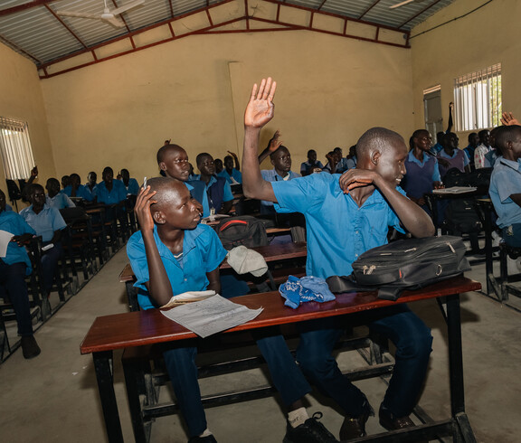 Viele jugendliche Schüler sitzen in einem Klassenraum auf Schulbänken. In der vordersten Reihe hebt ein Junge die Hand.