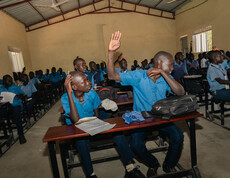 Viele jugendliche Schüler sitzen in einem Klassenraum auf Schulbänken. In der vordersten Reihe hebt ein Junge die Hand.