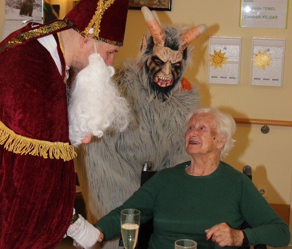 Nikolaus reicht einer Bewohnerin im Rollstuhl die Hand, im Hintergrund steht der Krampus.