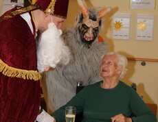 Nikolaus reicht einer Bewohnerin im Rollstuhl die Hand, im Hintergrund steht der Krampus.