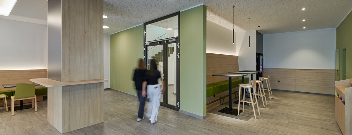 Innenansicht Aula Caritas Schule Wiener Neustadt, 2 Mädchen gehen zur Glastür Richtung Stiegenhaus