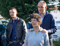 Ein junger Mann mit geistiger Beeinträchtigung steht lächelnd im Vordergrund, begleitet von zwei männlichen Betreuern, die ihm unterstützend zur Seite stehen.