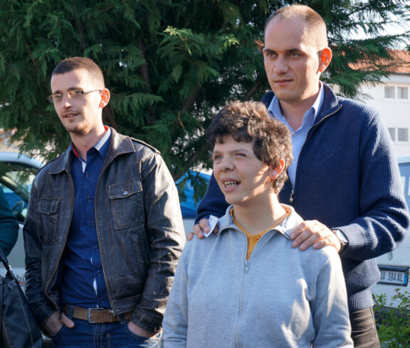 Ein junger Mann mit geistiger Beeinträchtigung steht lächelnd im Vordergrund, begleitet von zwei männlichen Betreuern, die ihm unterstützend zur Seite stehen.