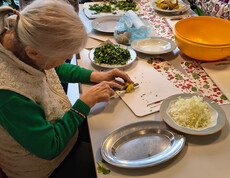 Eine Seniorin schneidet Gemüse, daneben liegen gehackte Kräuter, Zwiebeln und Kartoffeln bereit.