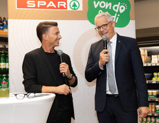 Zwei Personen sprechen mit Mikrofon vor SPAR-Logo, stehen vor Getränkeregal im Markt.