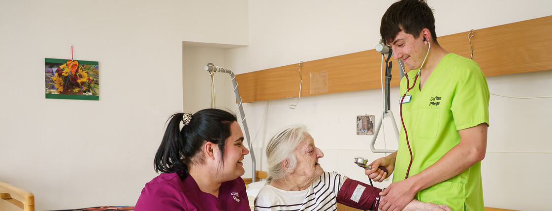 Zwei Pflegekräfte begleiten eine ältere Person beim Blutdruckmessen in einem Pflegezimmer der Caritas.