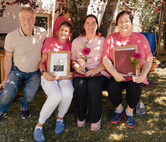 Vier Mitarbeitende sitzen im Garten und halten Urkunden und Blumen nach Ehrung beim Ratschnachmittag