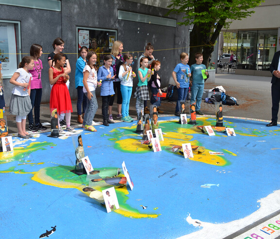 Bregenzer Bürgermeister Markus Lienhart steht mit Kindern im Volksschulalter im Freien. Sie schauen sich ein großes Kunststück an, das auf Asphalt ausgelegt worden ist. Es repräsentiert die Weltkugel und ist somit blau grün und braun. Auf den jeweiligen Kontinenten sind Kärtchen aufgestellt. Auf diesen sind Kinder zu sehen. 