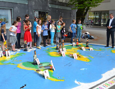 Bregenzer Bürgermeister Markus Lienhart steht mit Kindern im Volksschulalter im Freien. Sie schauen sich ein großes Kunststück an, das auf Asphalt ausgelegt worden ist. Es repräsentiert die Weltkugel und ist somit blau grün und braun. Auf den jeweiligen Kontinenten sind Kärtchen aufgestellt. Auf diesen sind Kinder zu sehen. 