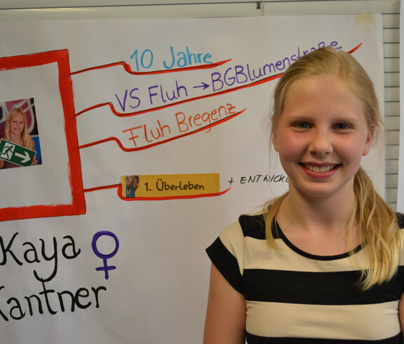 Eine Jugendliche mit blonden langen Haaren und gestreiften Oberteil steht vor einem Flipchart. Sie lächelt in die Kamera. 